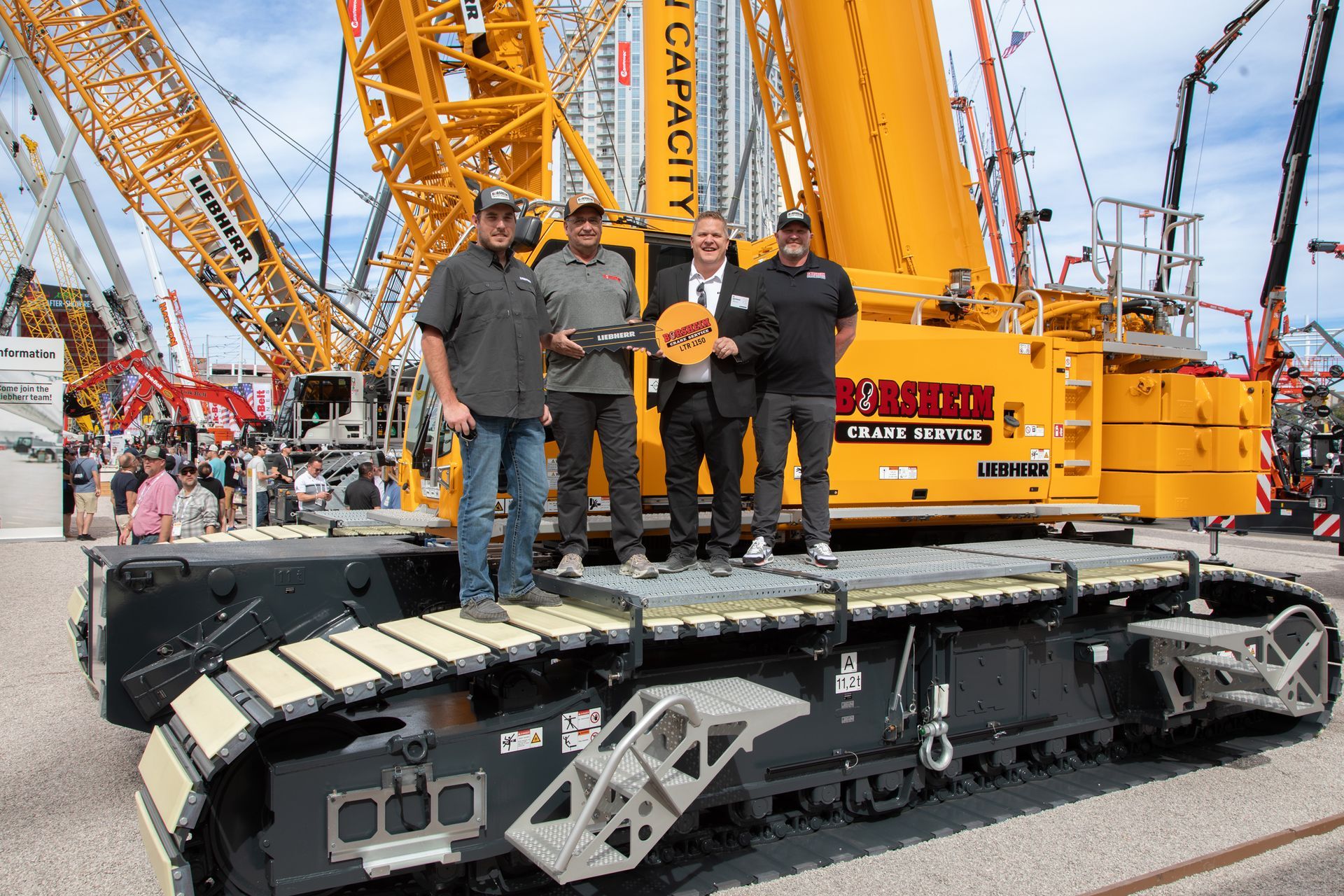 Four men standing on a large black tracked machine at a construction site with orange cranes in the background