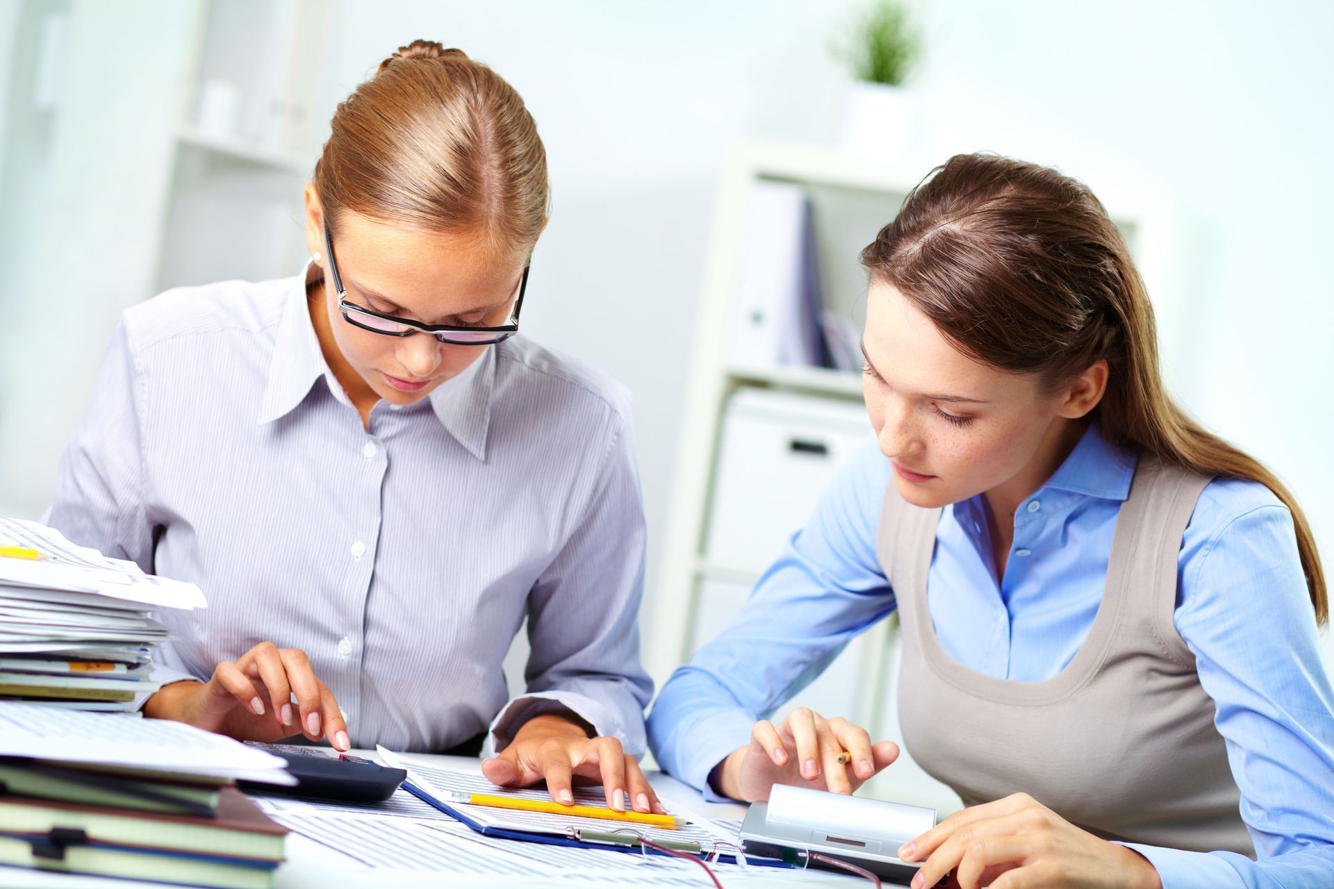 Two women reviewing documents, one using a calculator, in an office setting.