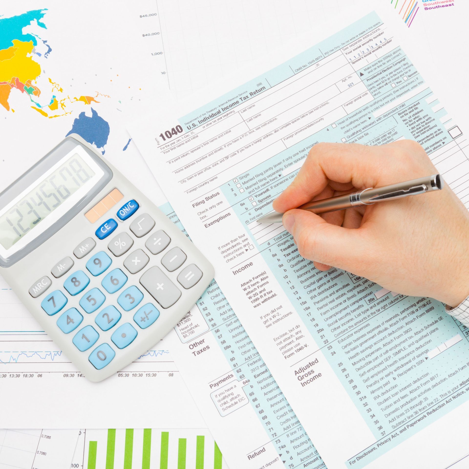 Hand filling out a 1040 tax form with a pen next to a calculator and financial graphs.