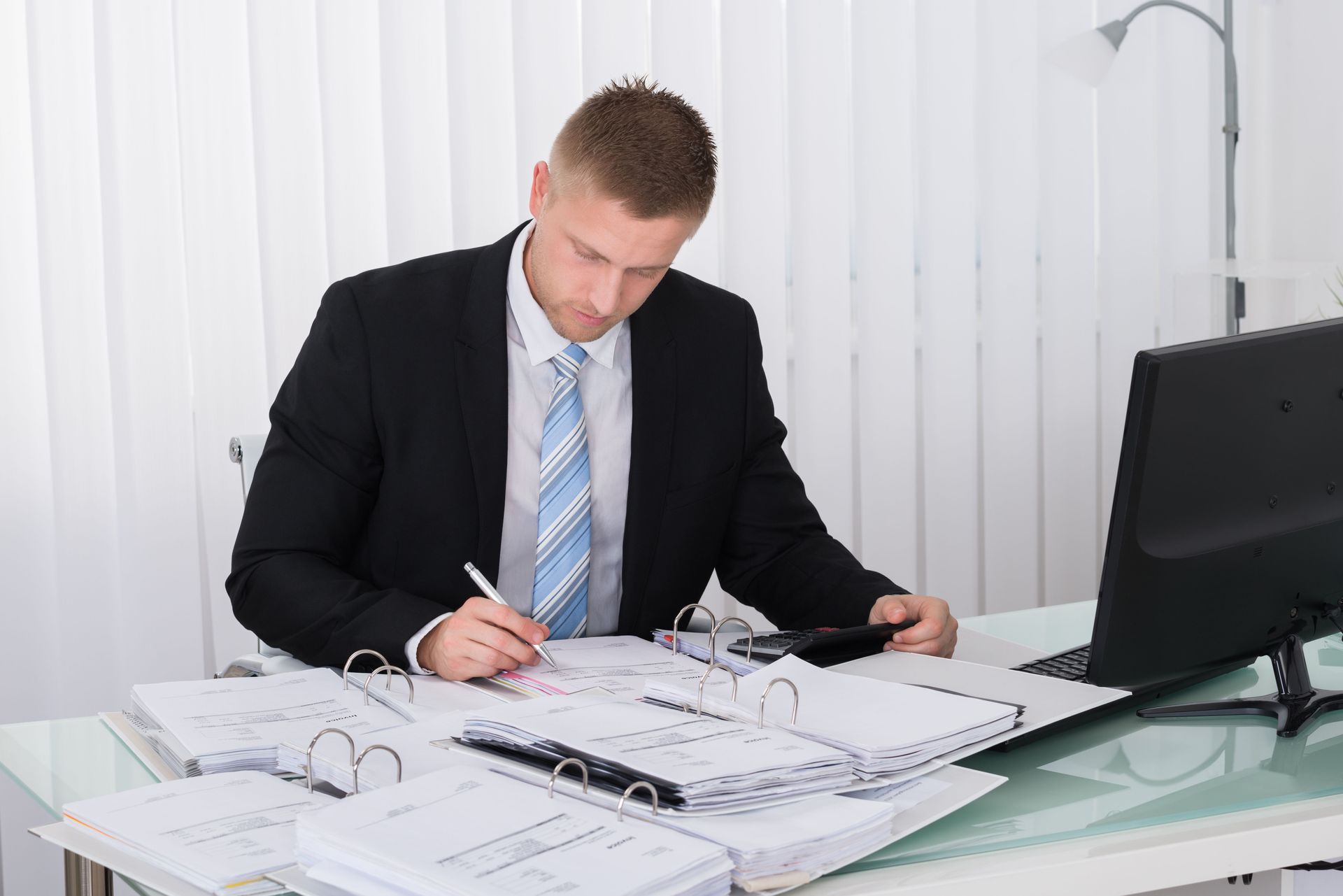 Man in suit reviews documents at desk, using pen and calculator in office setting.