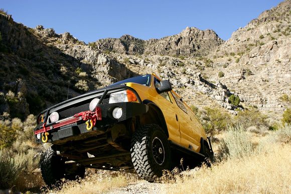 A yellow truck is driving down a dirt road with mountains in the background