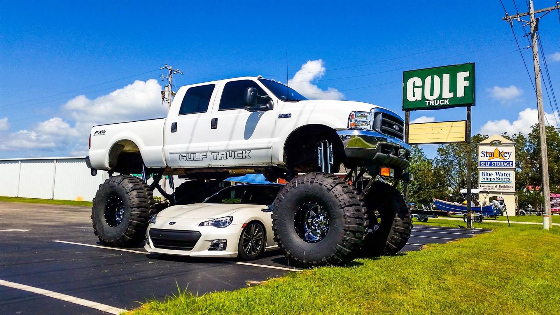 A white lifted pickup truck parked directly over a low-profile white sports car in a paved lot next to a golf sign.