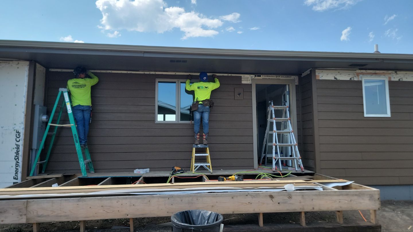Construction workers installing brown siding on a building under a blue sky.