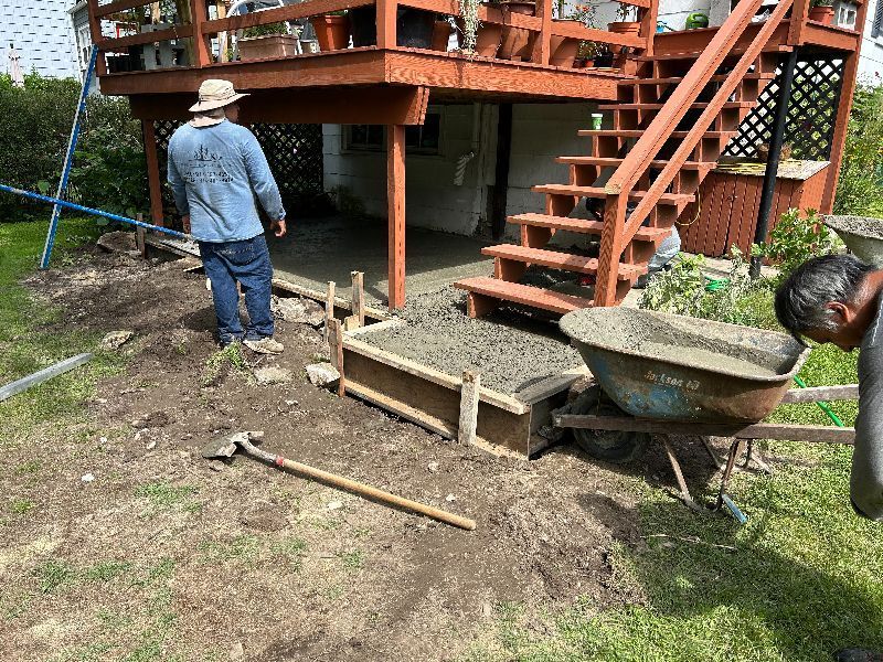 A man is pouring concrete into a wheelbarrow in front of a deck.