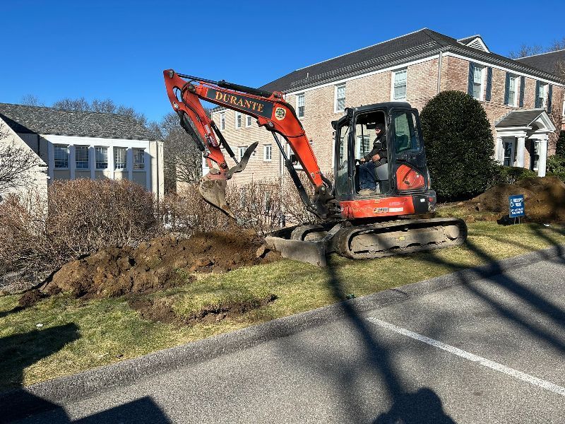 An excavator is digging a hole in the grass in front of a house.