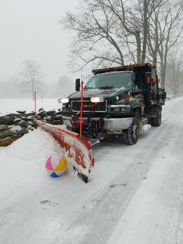 A snow plow is driving down a snowy road next to a beach ball.