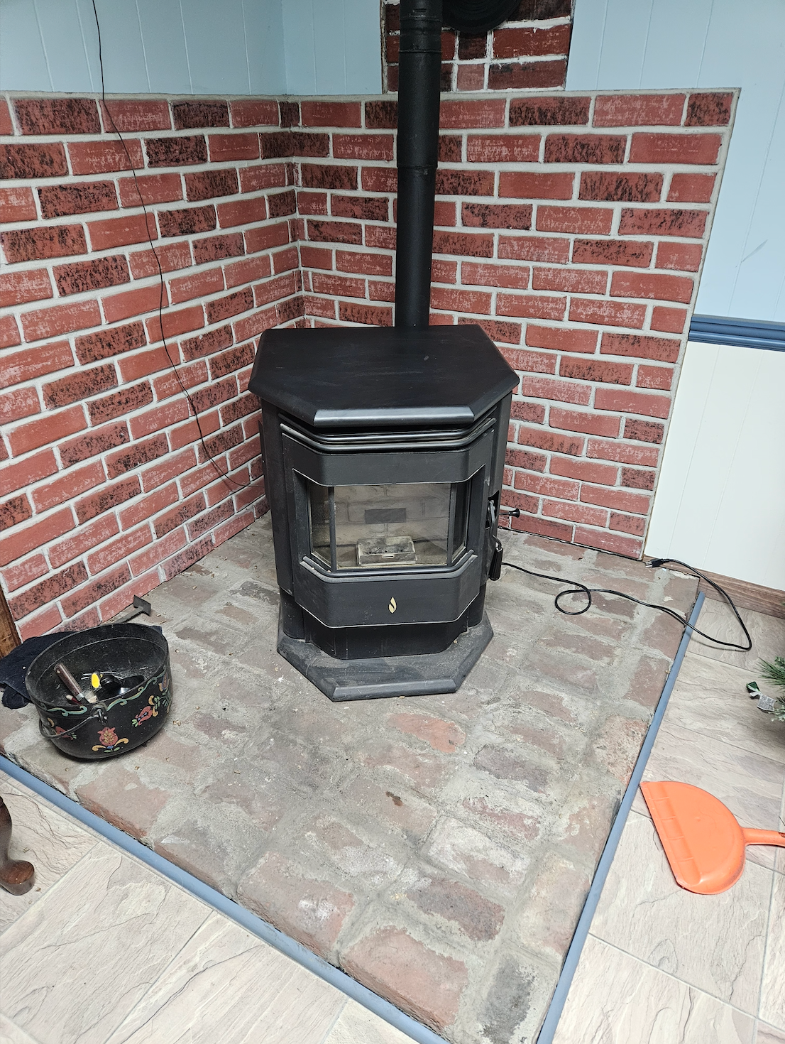Black wood stove in a brick corner on a tiled floor, with cleaning tools nearby.