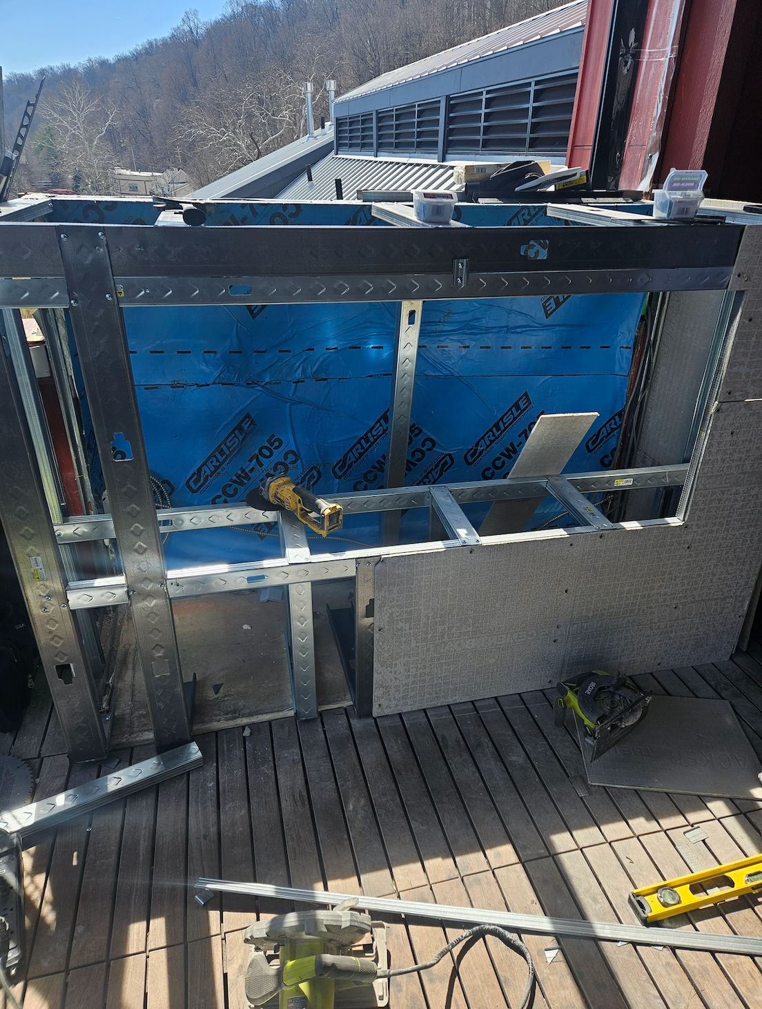 Metal-framed wall opening under construction with blue sheathing, on a wooden deck beside a building.