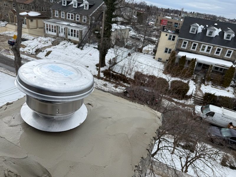 Rooftop metal vent on a flat roof, with snow-covered houses and bare trees in the background.