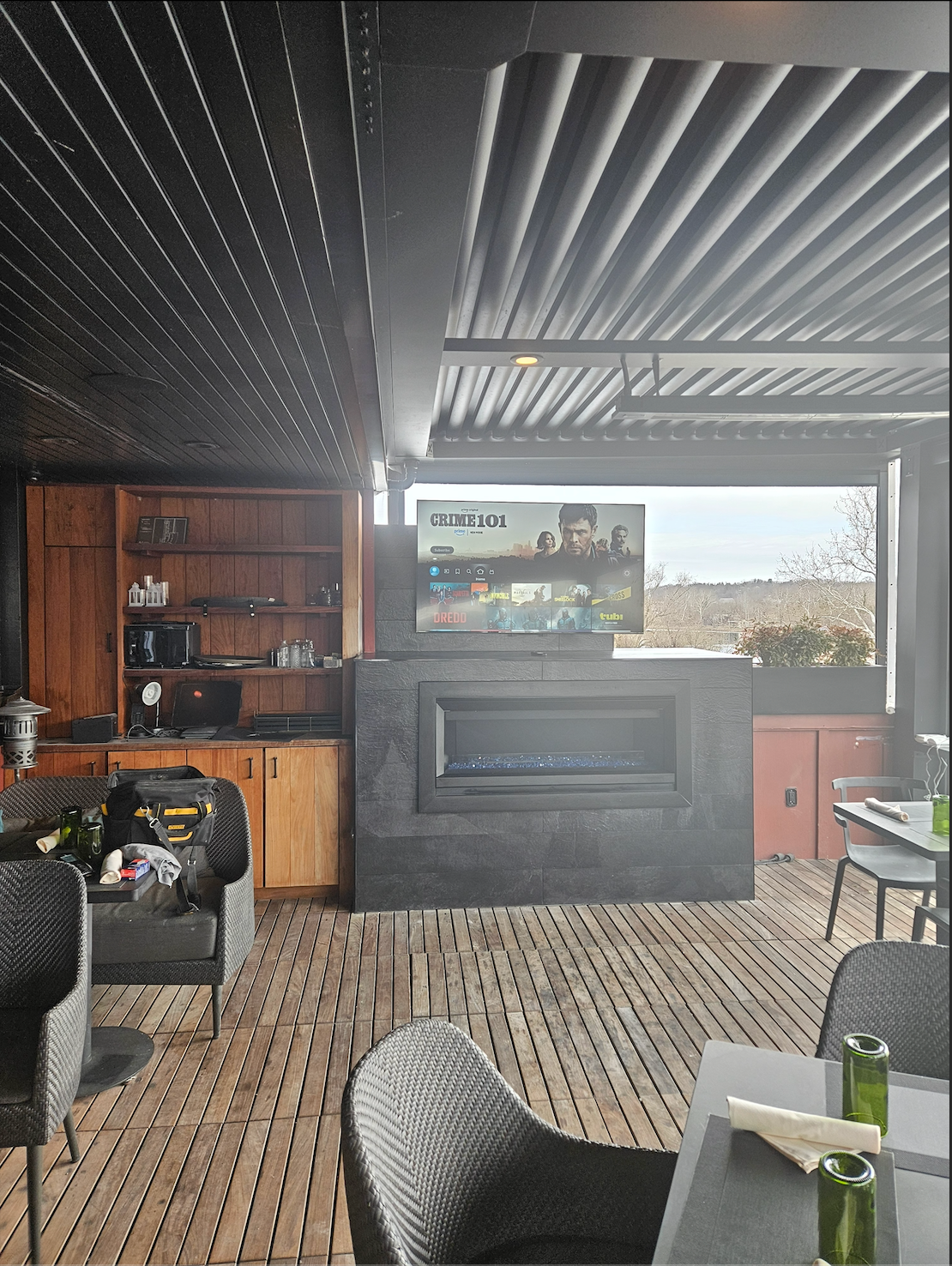 Modern patio lounge with black pergola ceiling, TV wall, wood shelves, and outdoor seating on a rug.