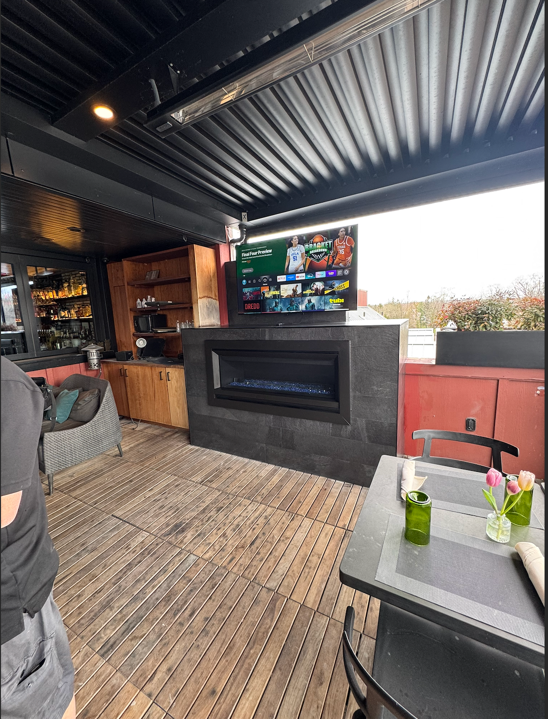 Open-air restaurant patio with TV, fireplace, wooden decor, and a table with drinks