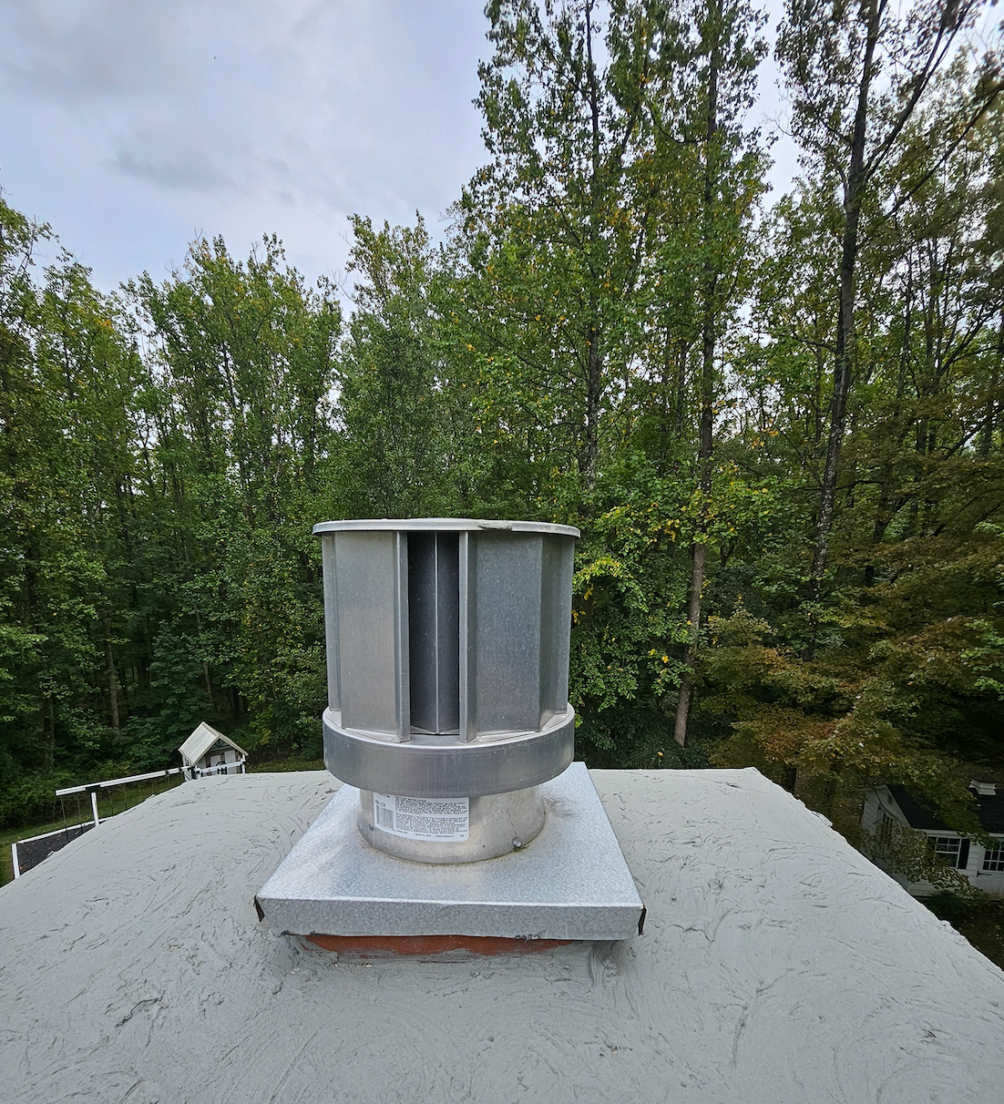 Rooftop metal vent with gray cap, surrounded by green trees under a cloudy sky