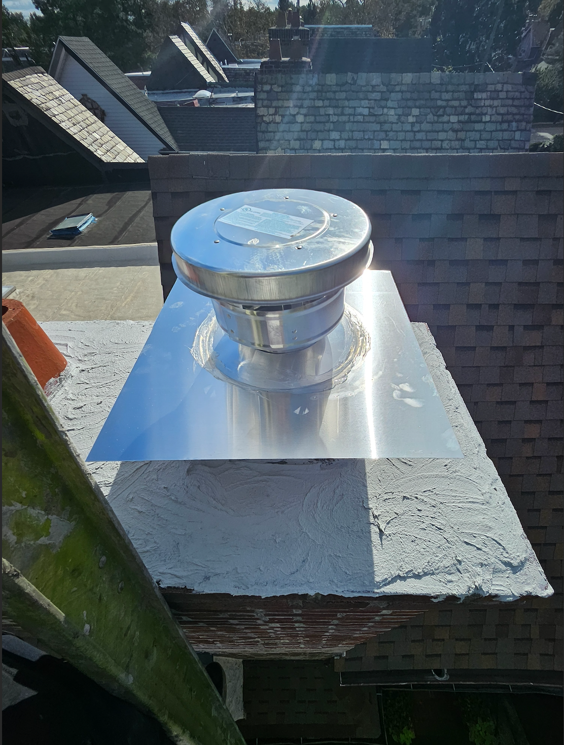 Metal roof vent cap on a shiny flashing base atop a white chimney against neighboring rooftops.