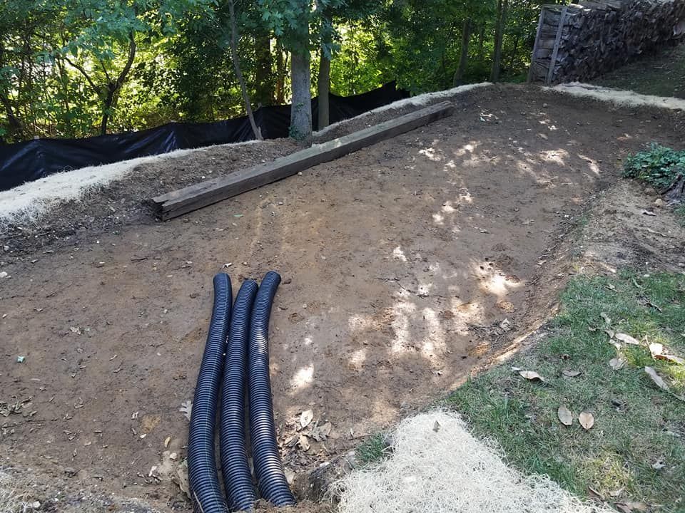A construction site with a flat dirt area, three drainage pipes, and timber beams, bordered by a fence and wood pile.