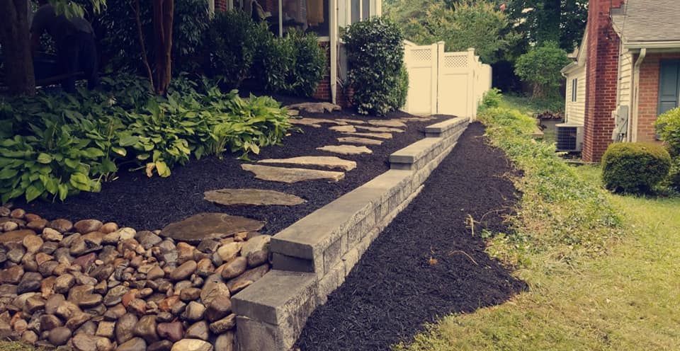 A landscaped yard featuring a stone block retaining wall, a stone walkway, dark mulch, and a large patch of river rocks.