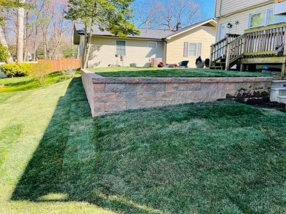 A residential backyard featuring a newly installed tan masonry retaining wall with fresh green sod.