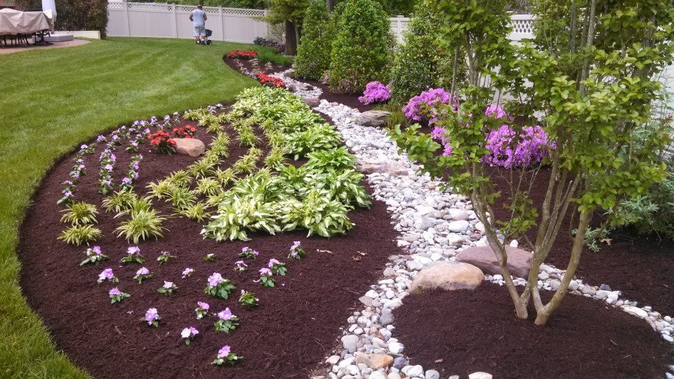 A landscaped garden bed with dark mulch, rows of hostas, purple flowers, and a curving stone path against a green lawn.
