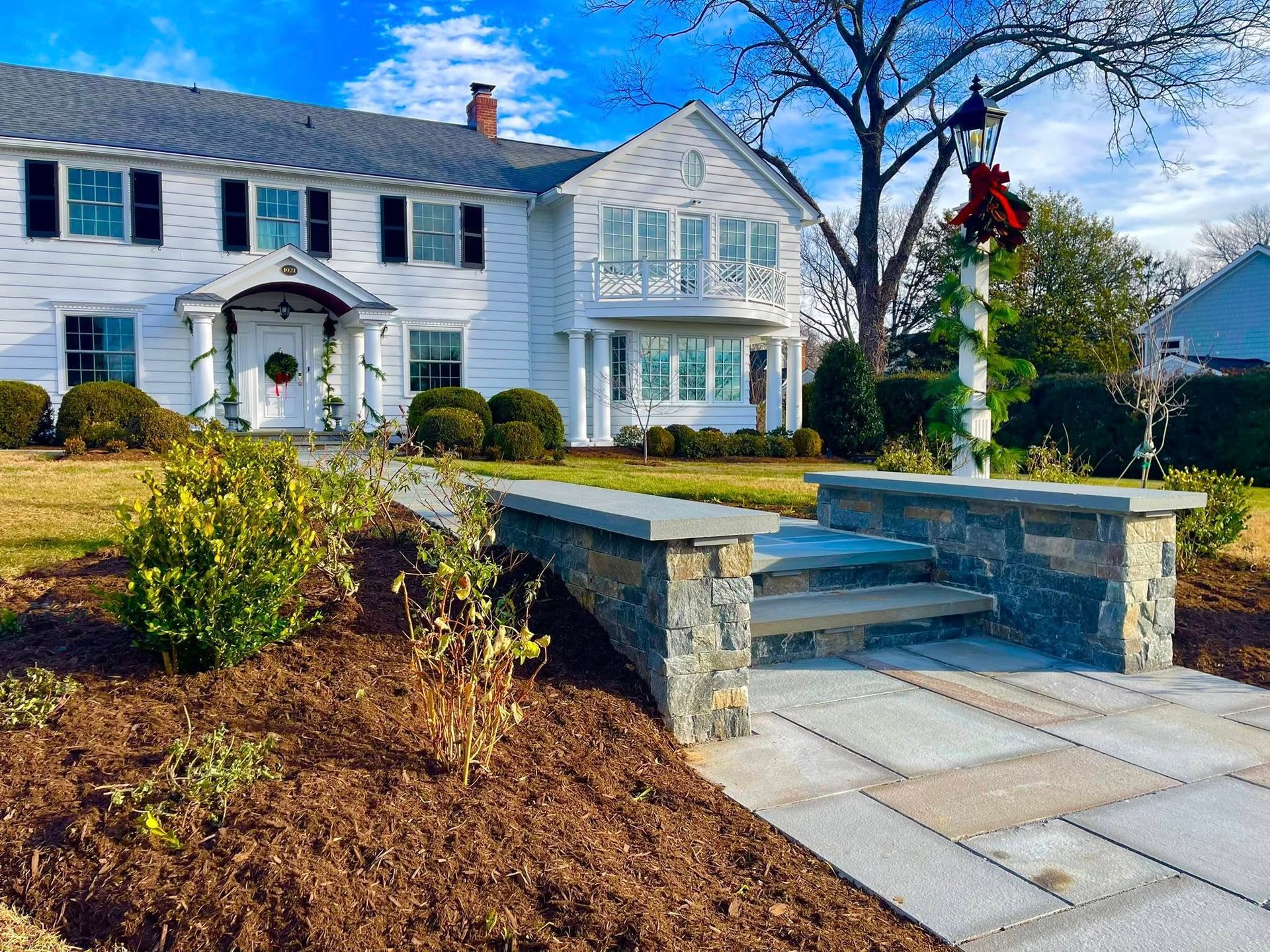 A classic white two-story colonial home with a stone walkway, landscaped garden, and decorated lamppost under blue skies.