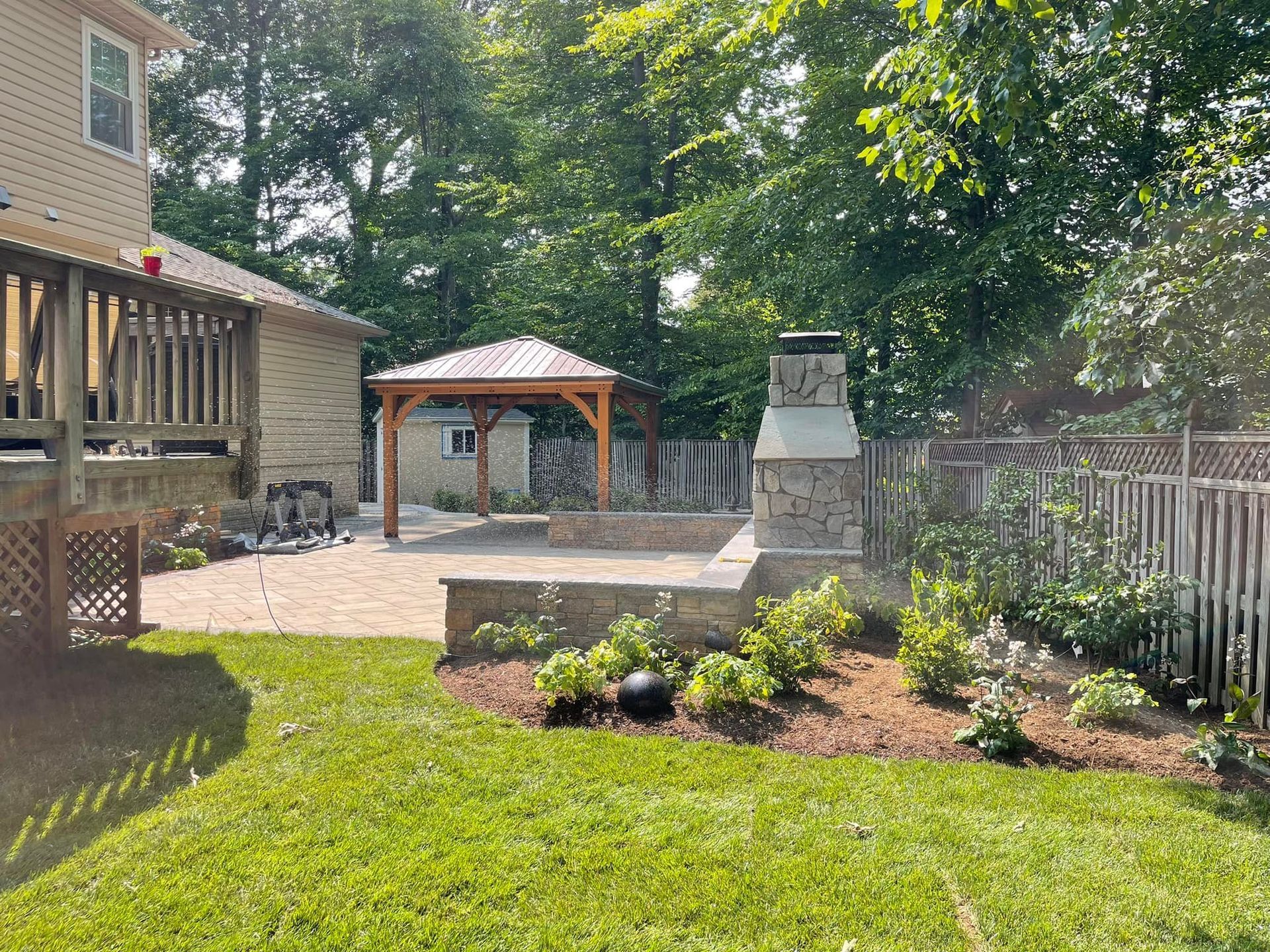 A sunny backyard featuring a stone patio, a wooden gazebo, an outdoor fireplace, and a house deck.