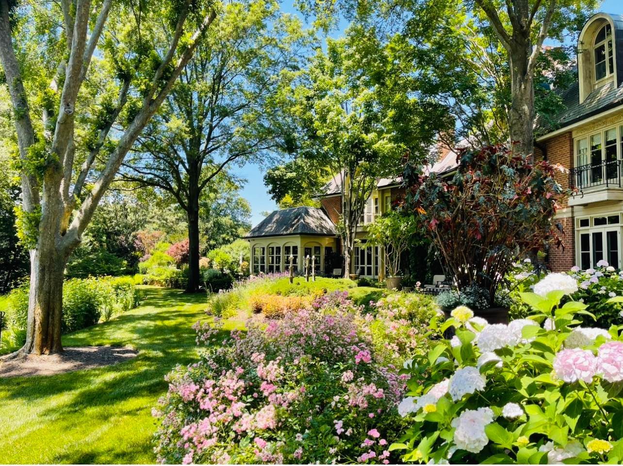 A lush garden with pink and white hydrangea bushes in the foreground leads to a multi-story home set among large trees.