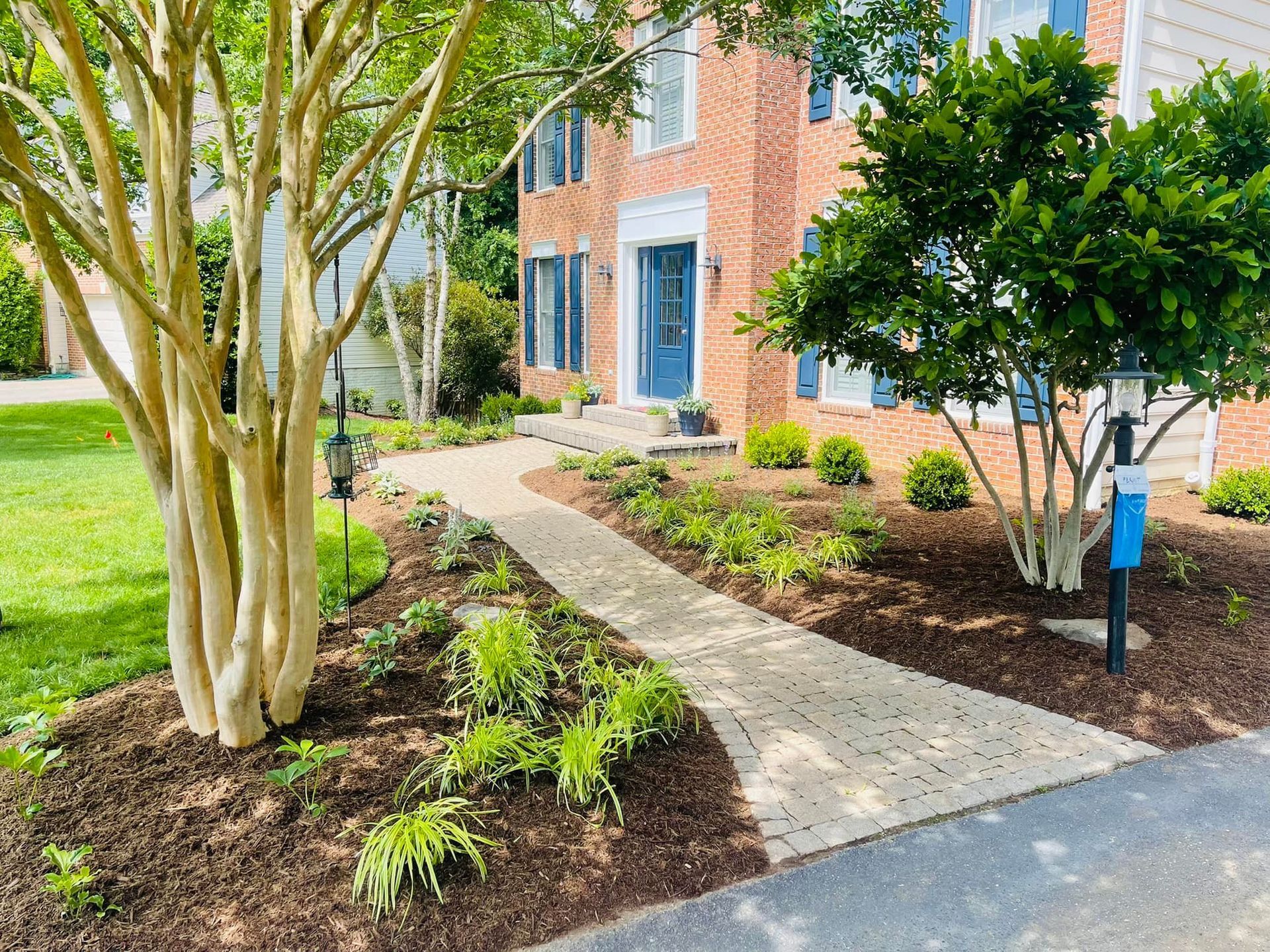 A paved walkway leads to the front door of a brick house, flanked by trees and garden beds with mulch and greenery.