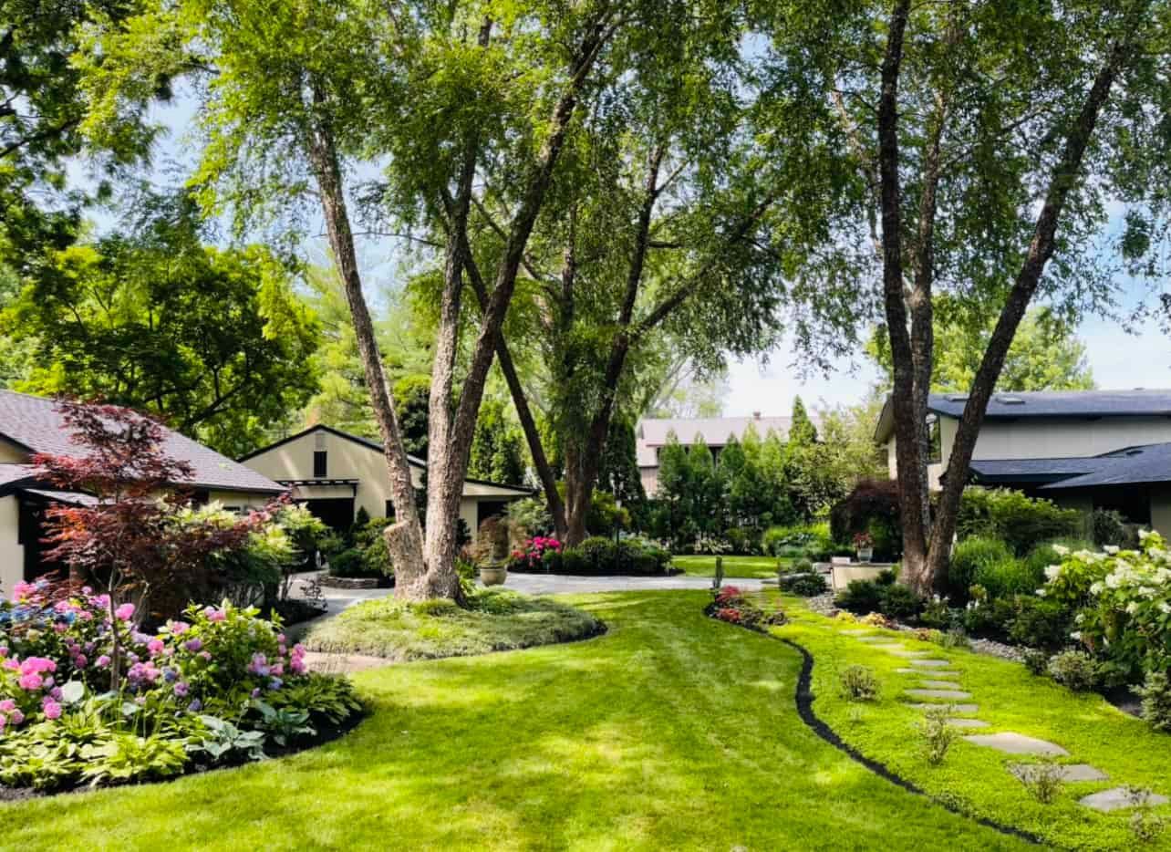A lush green backyard with manicured grass, large trees, a stone pathway, and vibrant garden beds surrounding a home.