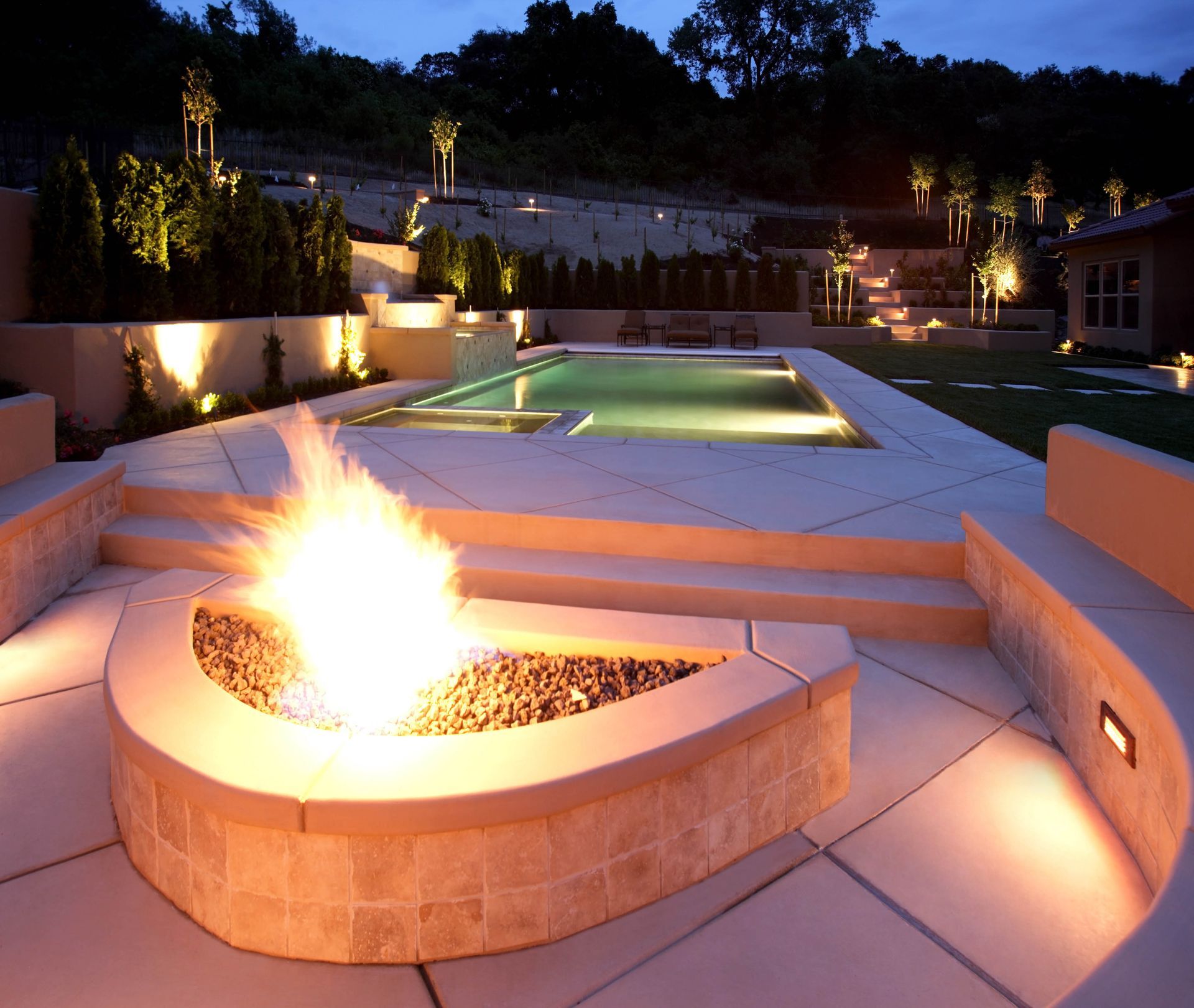 A lit fire pit in the foreground overlooks a modern illuminated swimming pool at night.