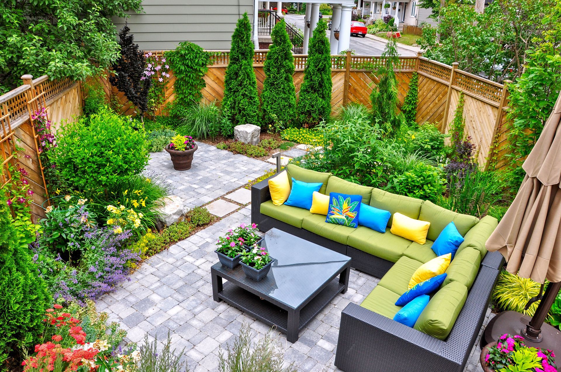 An outdoor patio featuring an L-shaped wicker sofa with green cushions and colorful pillows, set on grey stone pavers.