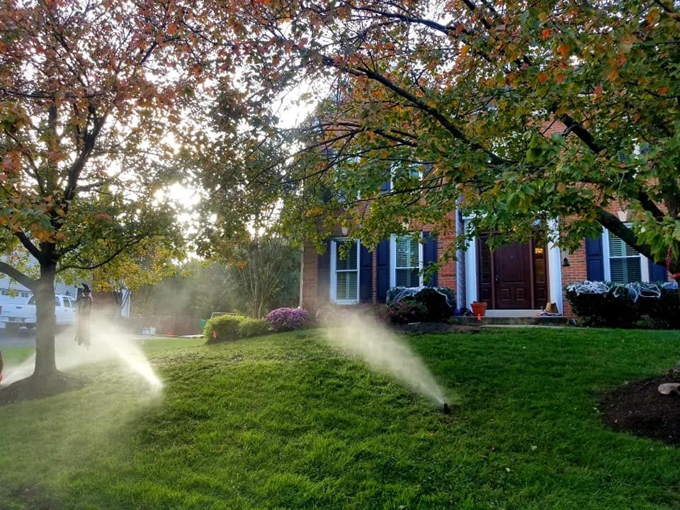 A two-story brick house with a lawn being watered by multiple sprinklers on a sunny autumn day.