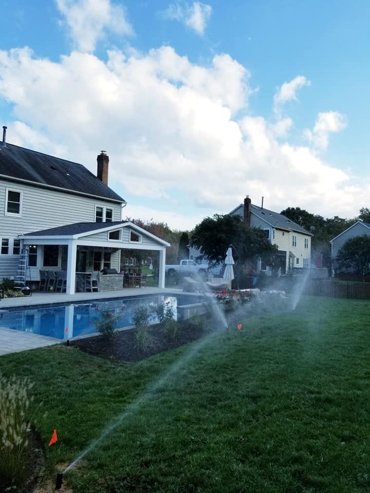 A lawn sprinkler sprays water across a grassy backyard with a swimming pool and patio next to a two-story house.