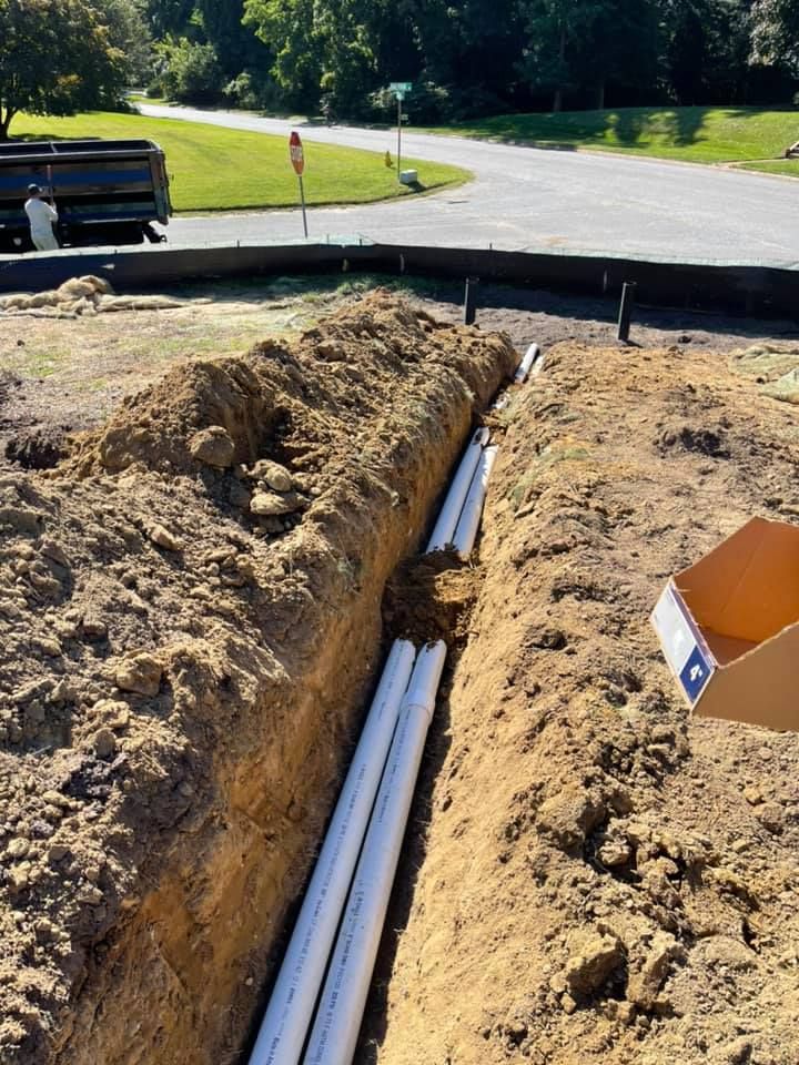 A construction trench containing several parallel white PVC pipes, with a truck and road visible in the background.