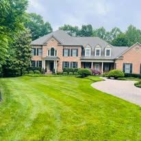 Large brick suburban home with a manicured lawn, lush green trees, and a circular driveway under a cloudy sky.