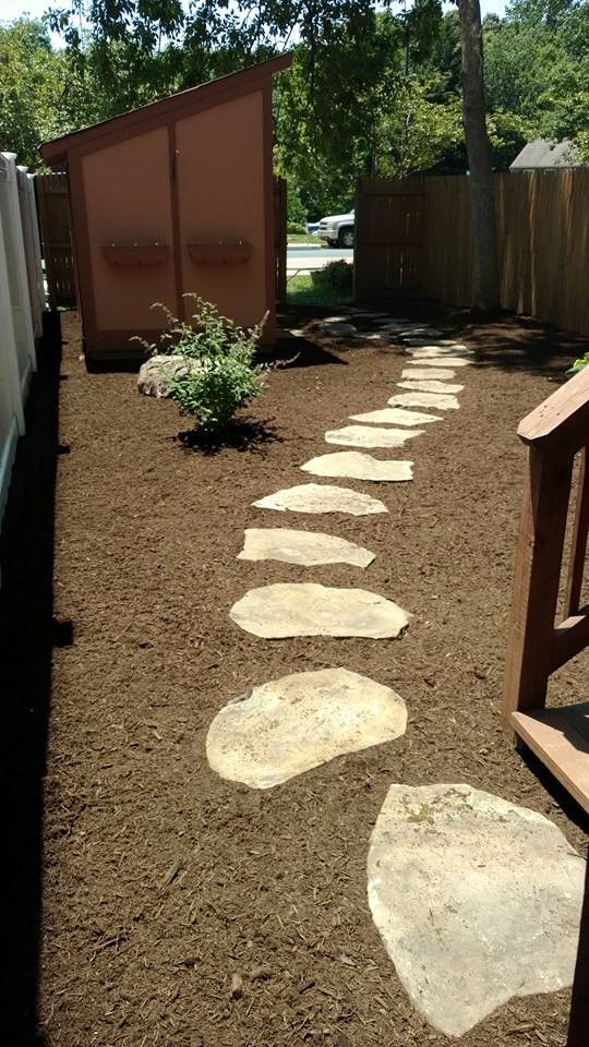 A winding stone path leads through a mulched yard toward a shed near a wooden fence and stairs.