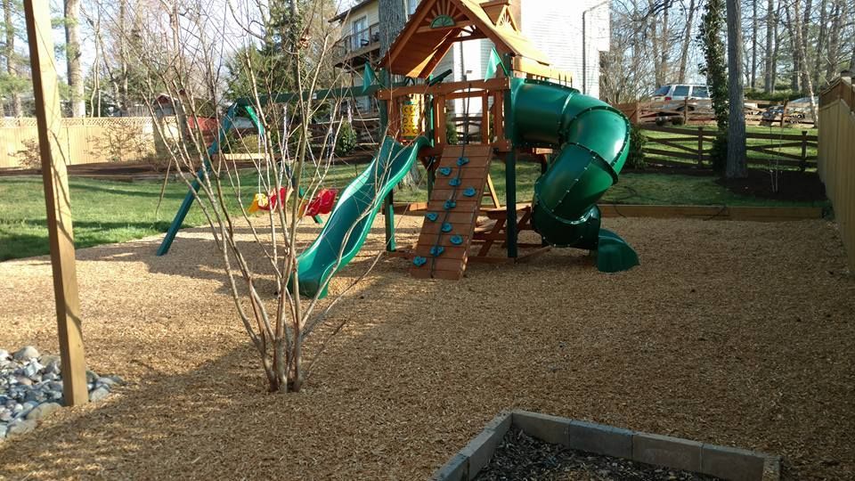 A wooden backyard playground set with a green slide, tube slide, and climbing wall on a wood-mulched surface.