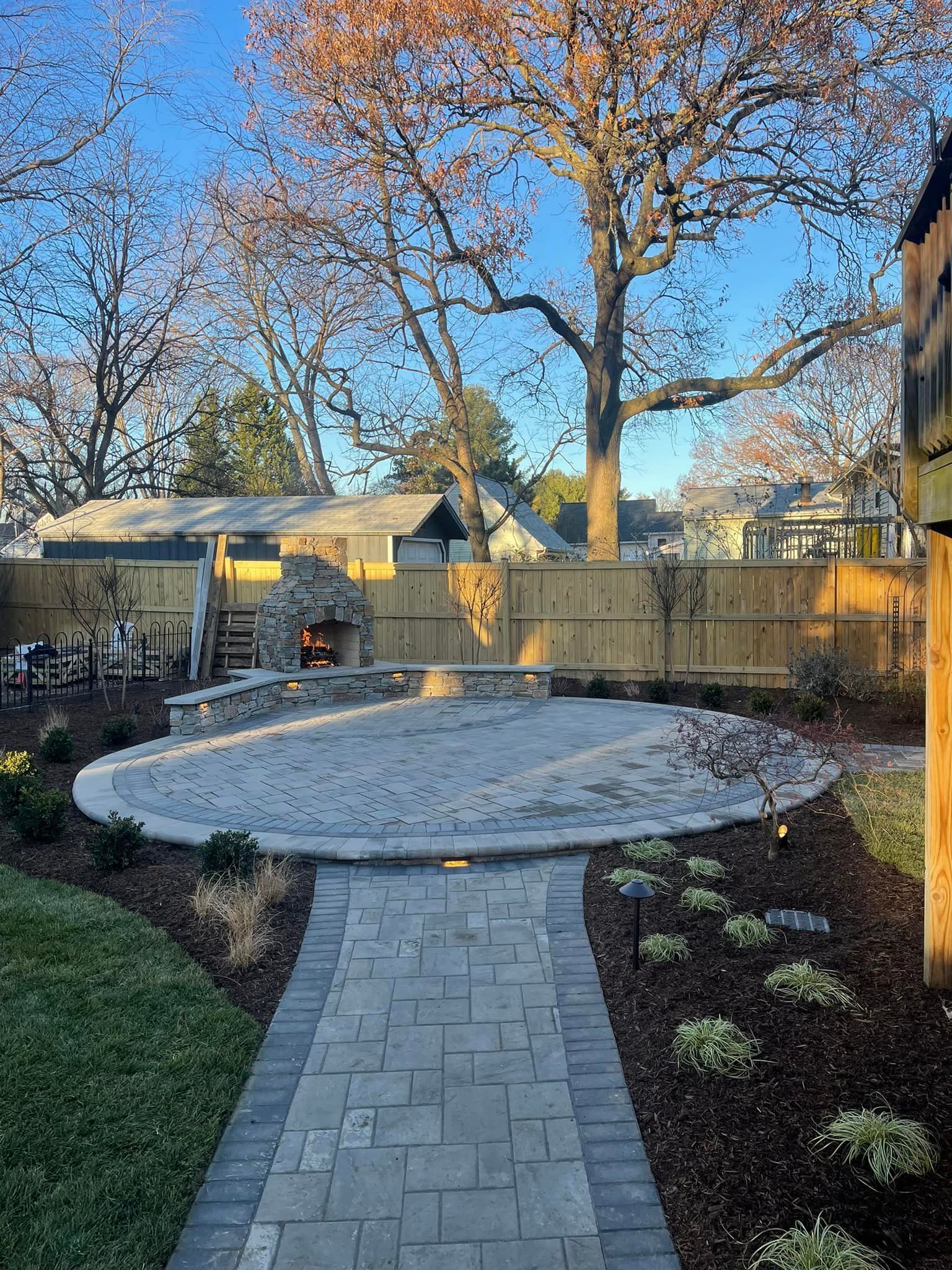 A stone patio with a built-in fireplace, connected to a paved walkway in a backyard with a wooden fence and large trees.