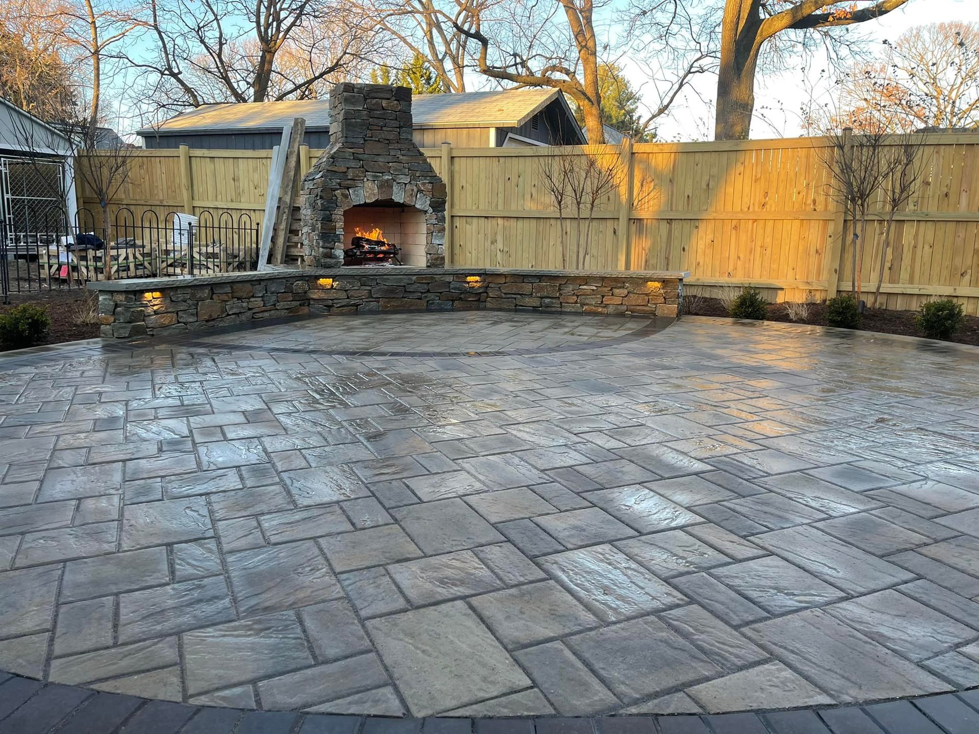 A stone-paved patio with a curved seating wall and a lit outdoor stone fireplace in front of a wooden fence.