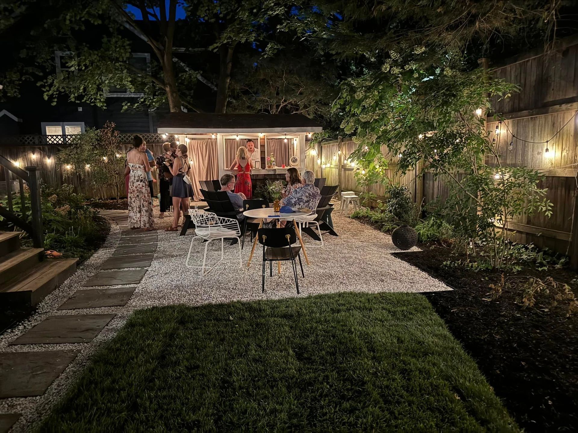 People gather for a social event in a backyard at night, illuminated by string lights, with chairs on a gravel patio.