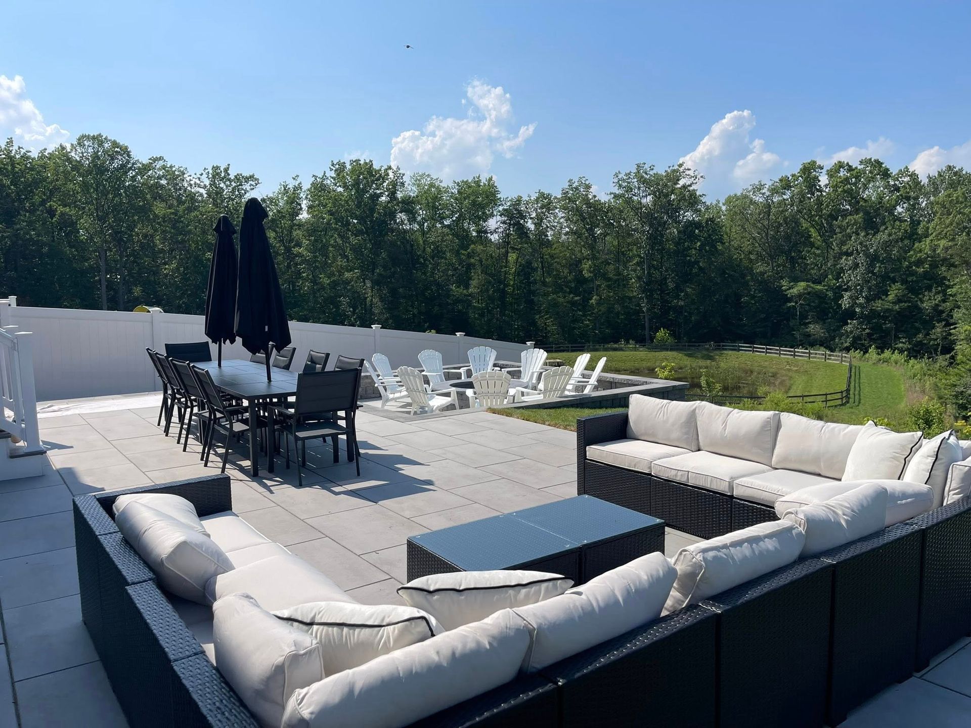 A large outdoor patio with dark wicker sofa seating, a dining set with black umbrellas, and chairs facing a wood line.