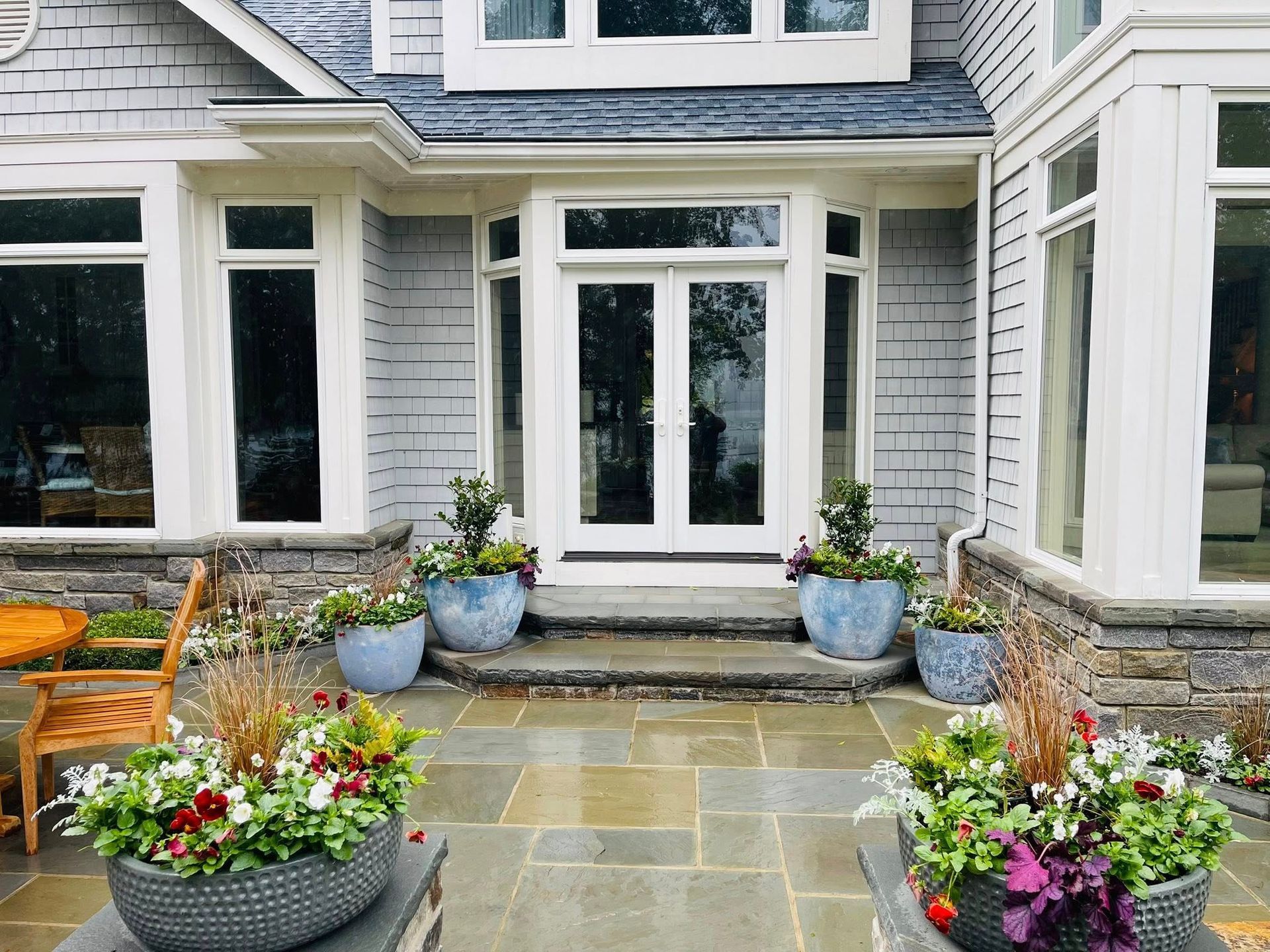 A stone patio with potted flowers in front of a house entrance featuring French doors and grey shingle siding.