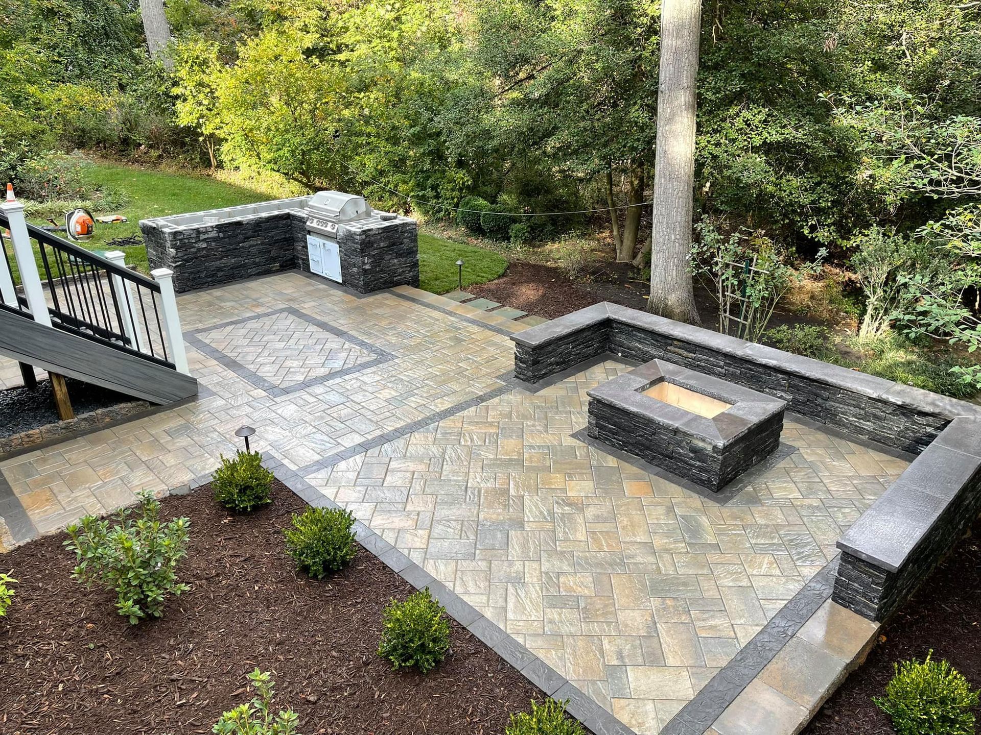 A high-angle view of a backyard patio featuring paver stones, a square fire pit, built-in stone seating, and a staircase.