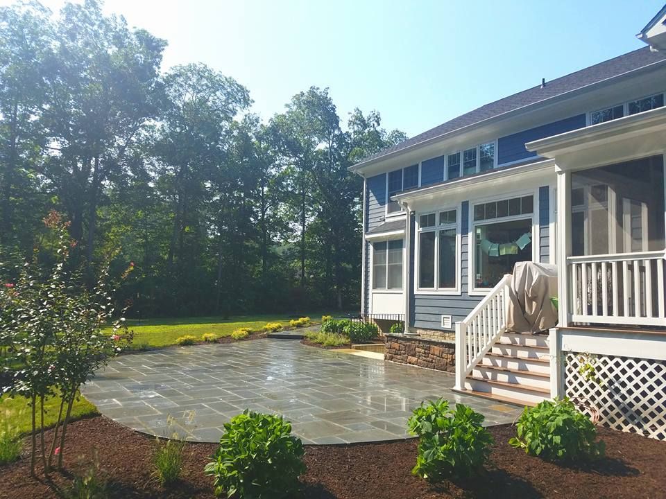 A backyard with a stone patio, a small staircase leading to a blue house, surrounded by green trees and mulch beds.