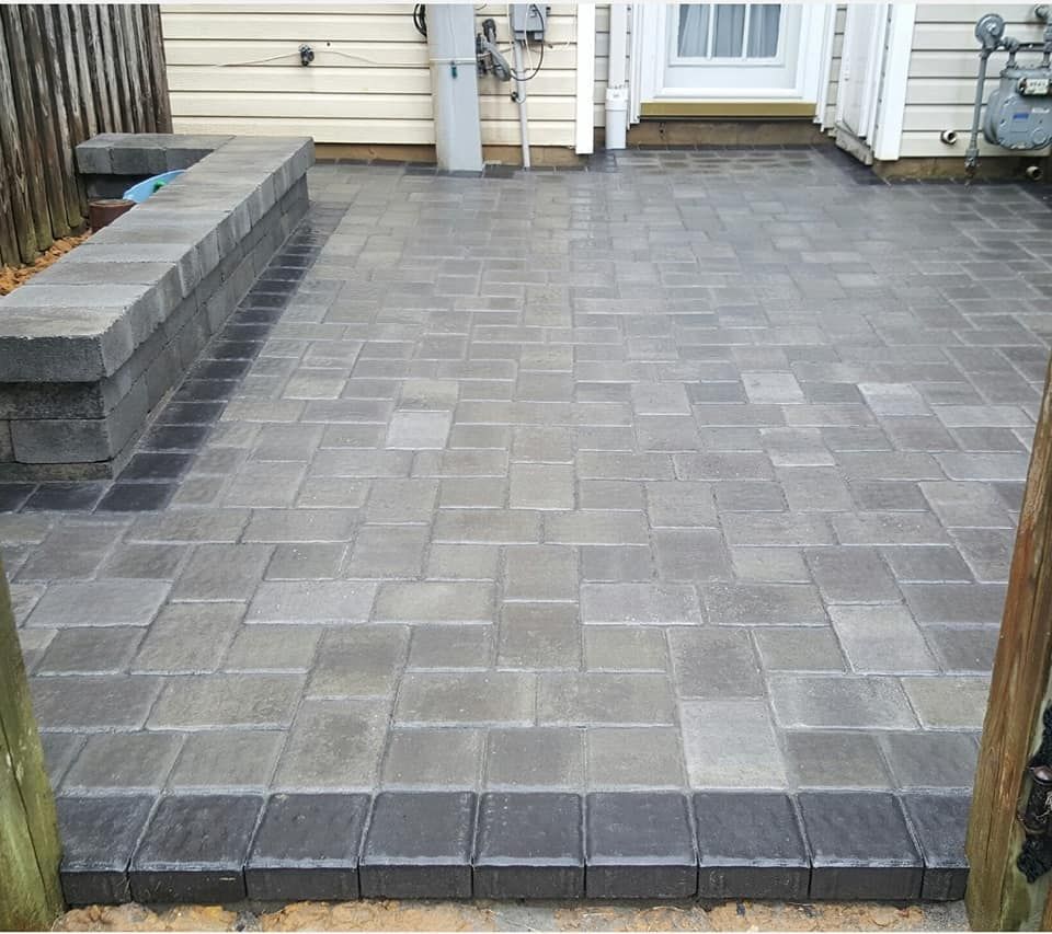 A stone patio with a gray paver floor, a matching stone built-in bench, and a dark border, set against a house exterior.