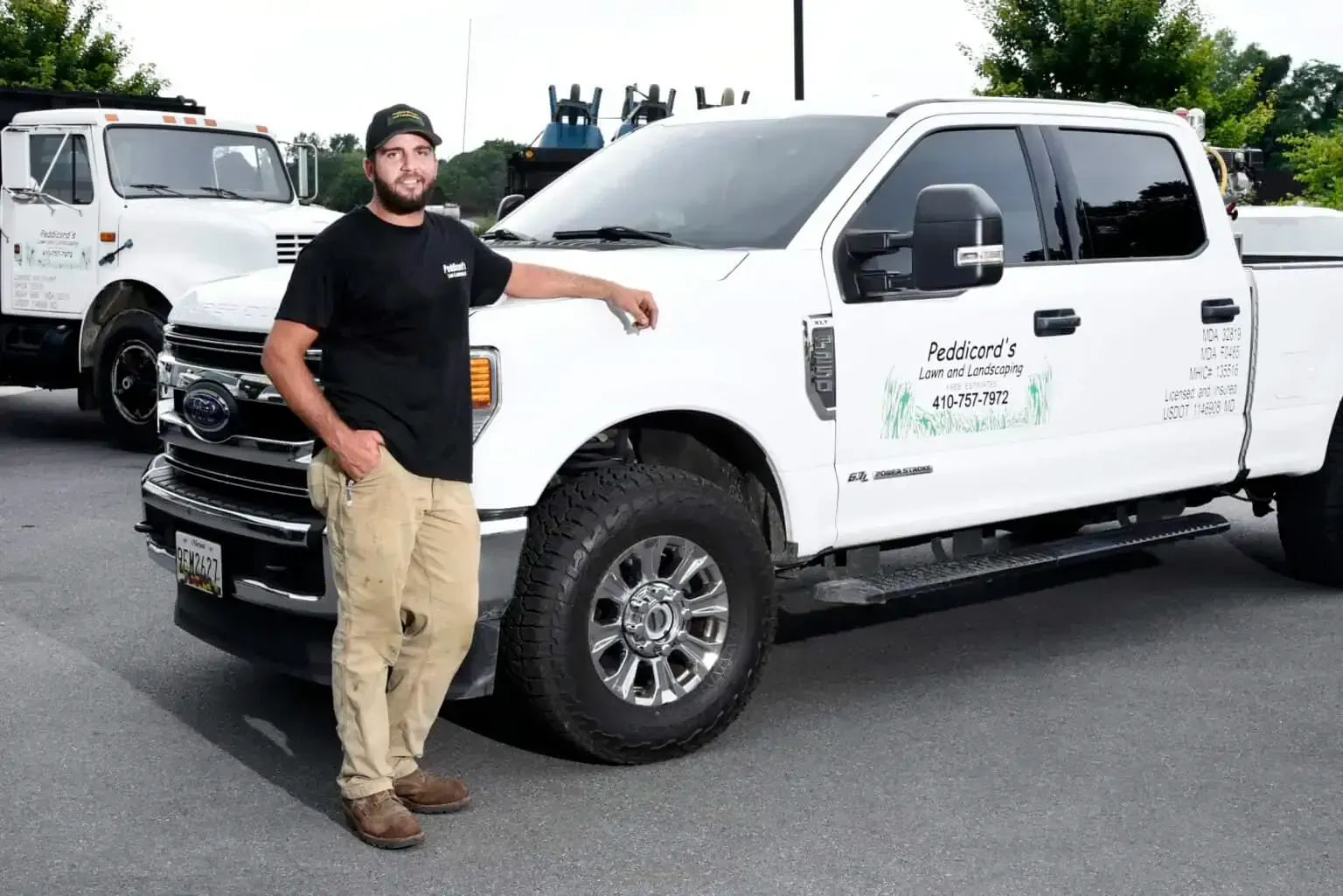 A smiling person wearing a black t-shirt and tan pants standing next to a white pickup truck in an outdoor lot.