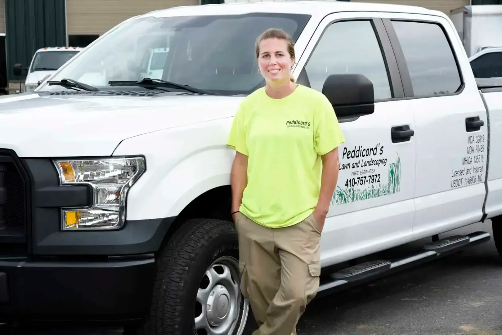 A person in a bright yellow shirt and khaki pants leans against a white pickup truck with logo branding on the side.