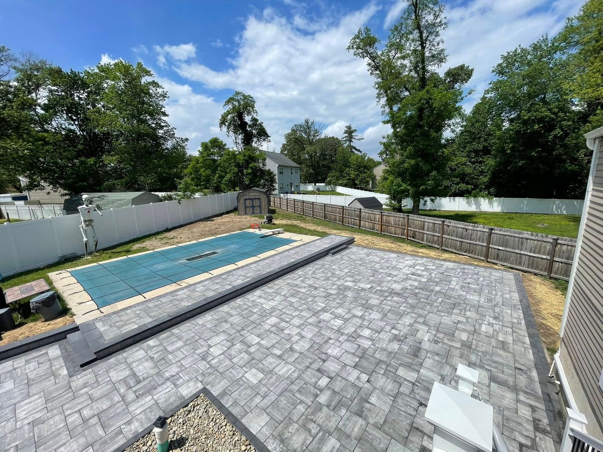 A backyard with a large grey paver patio, an in-ground pool covered with a blue tarp, and white vinyl perimeter fencing.