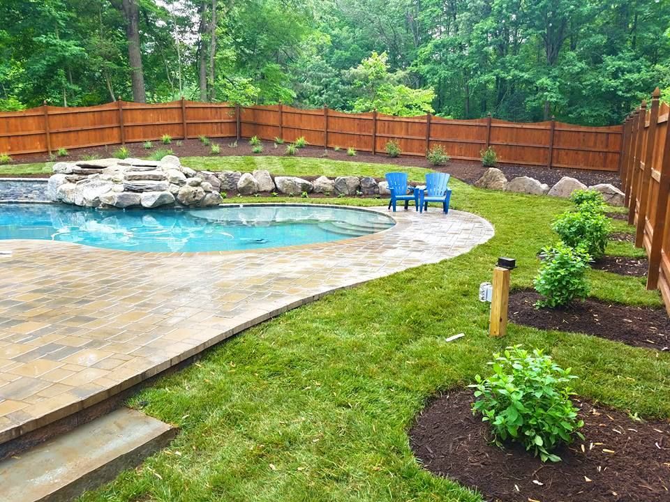 A backyard with an in-ground pool, stone water feature, paved patio, wooden privacy fence, and manicured garden beds.