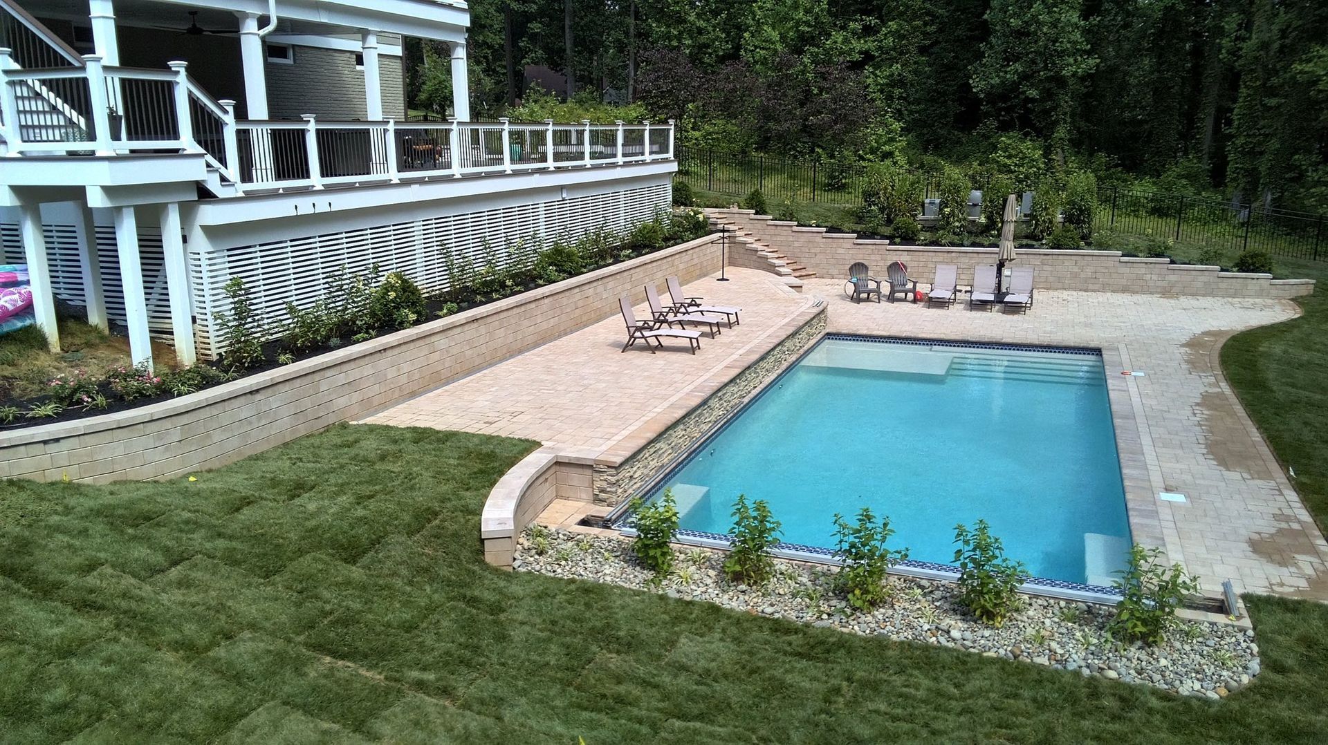 A backyard features a rectangular swimming pool next to a patio with lounge chairs, below a raised deck attached to a home.