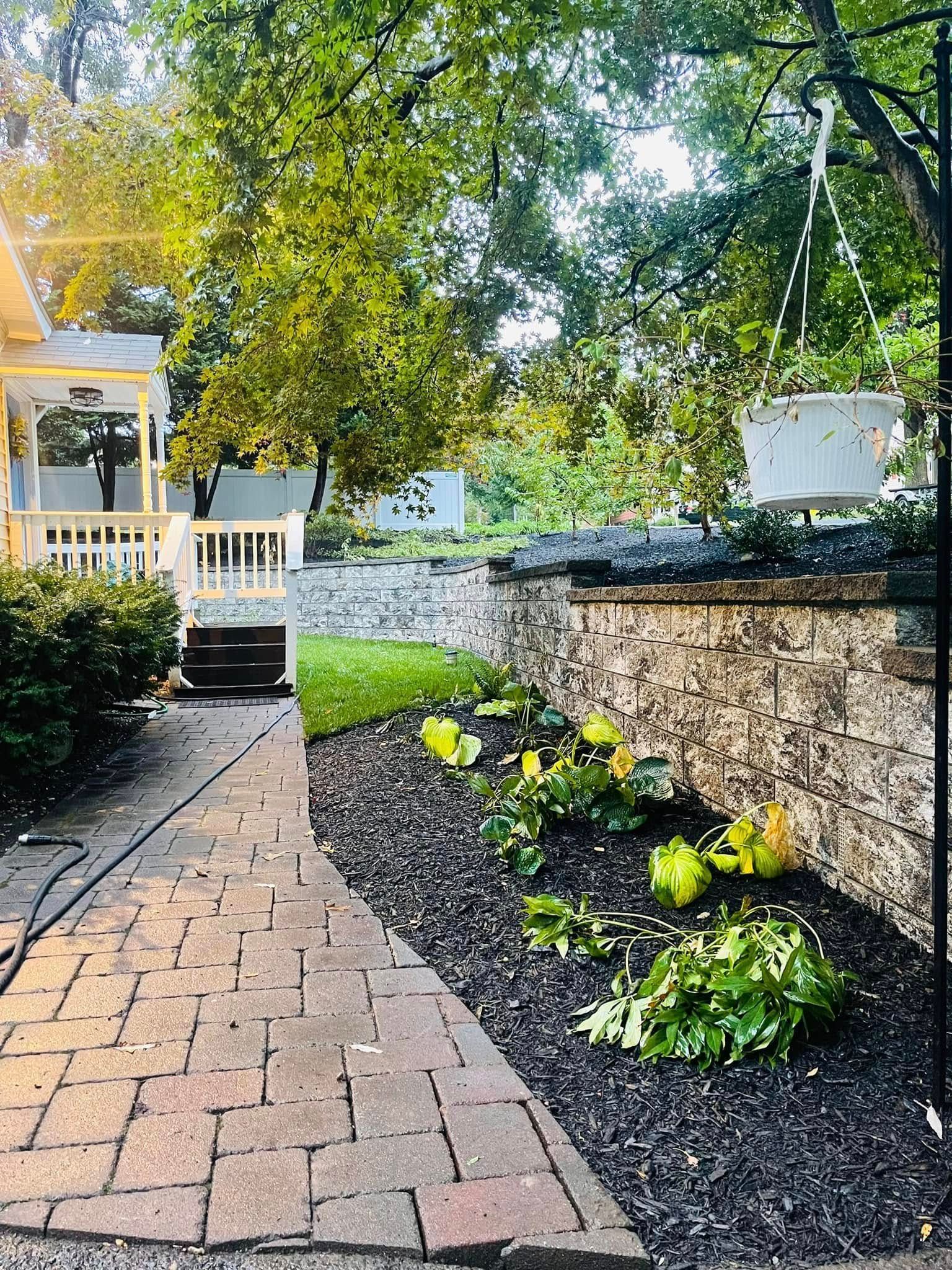 A brick walkway leads toward a house porch beside a stone retaining wall and dark mulch flower beds.