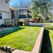 A house with a screen porch and wooden deck overlooks a grassy backyard enclosed by a tan stone retaining wall.