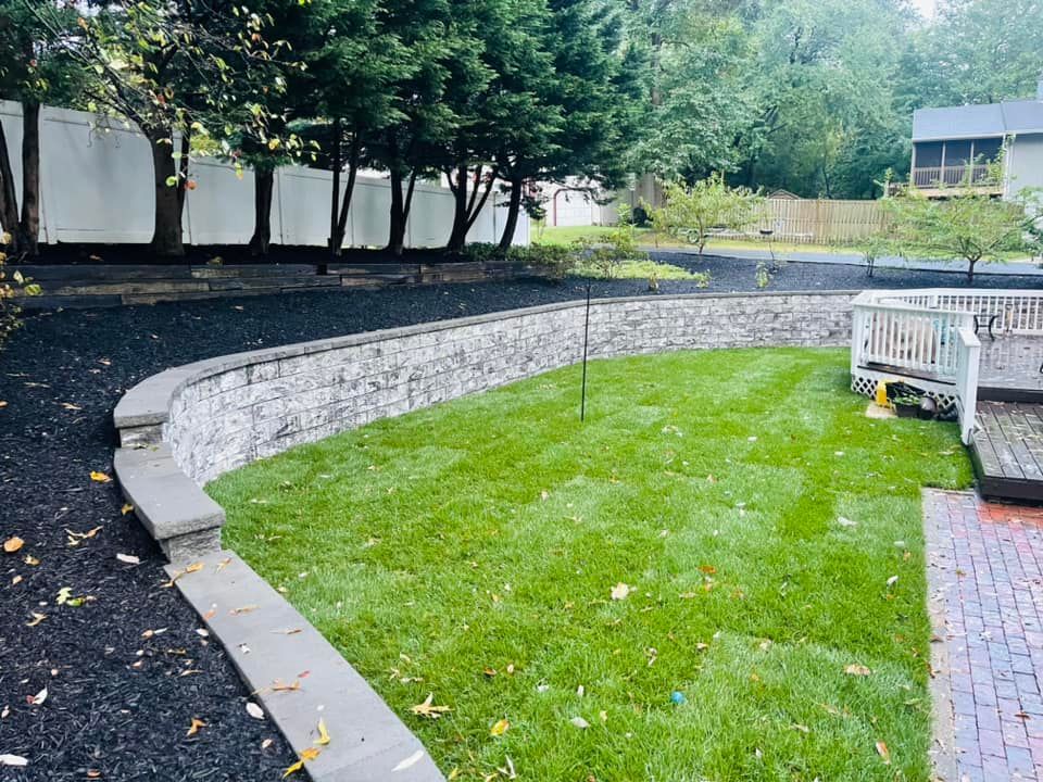 A landscaped backyard featuring a curved stone retaining wall, fresh green grass, and beds of dark mulch under tall trees.